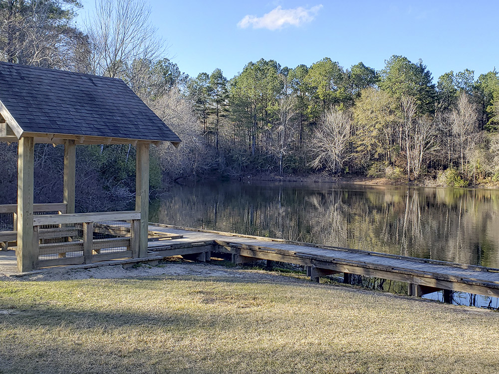 small pavilion and fishing pier extend over still pond surrounded by trees 