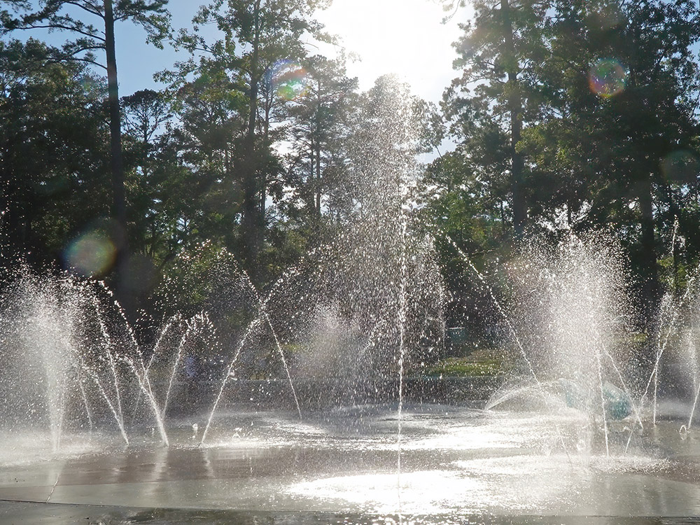water fountains on splash pad with trees in background backlit by sun in Jimmie Davis State Park