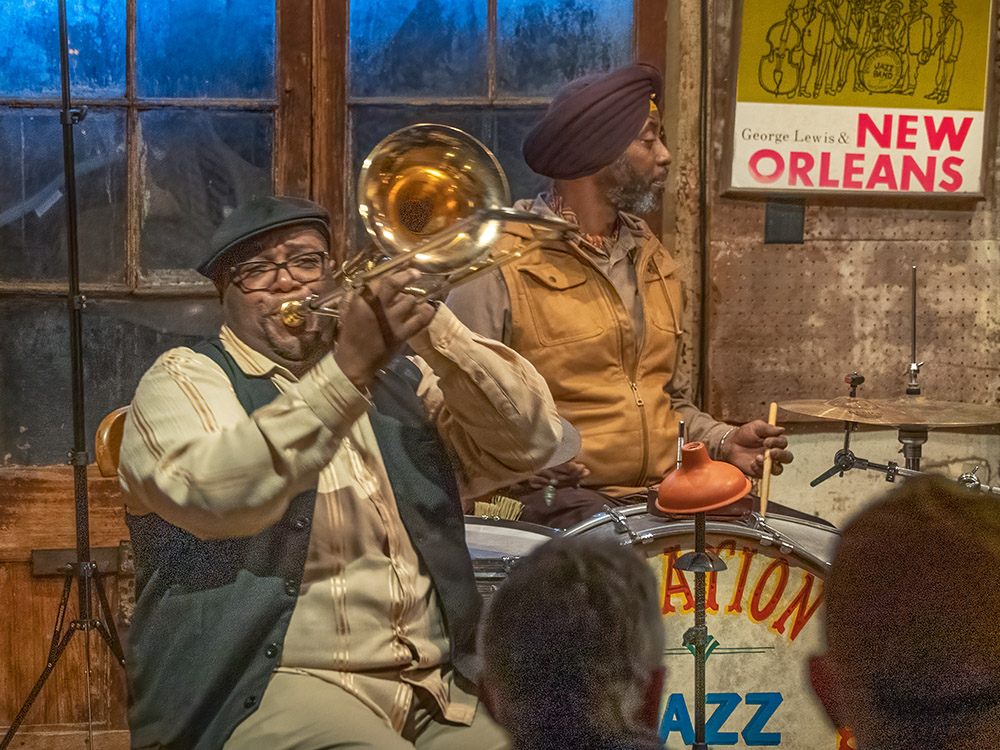 trombone player wearing cap and vest in front of drummer at Preservation Hall
