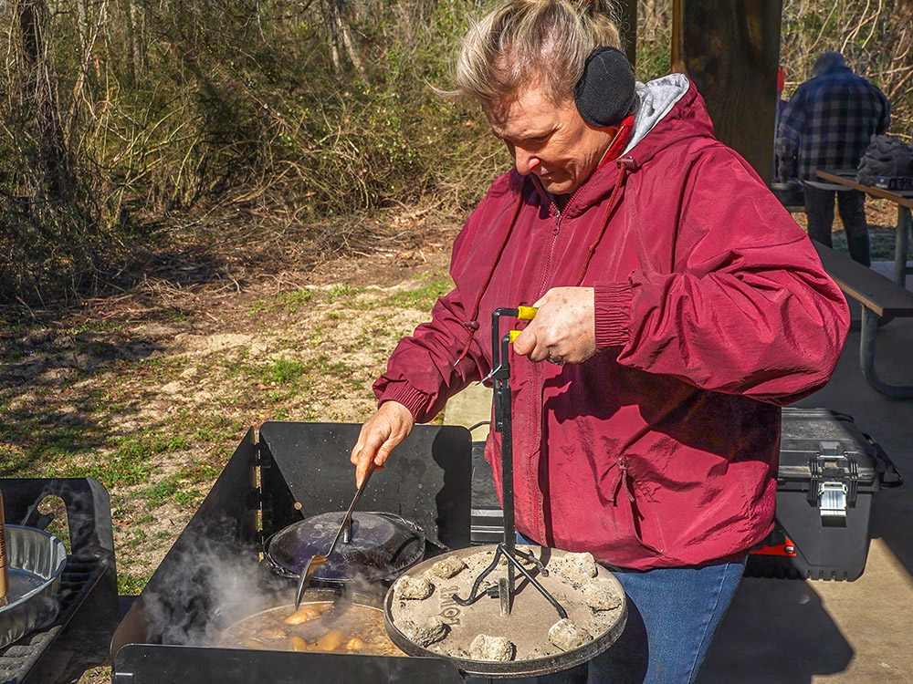 woman in red jacket stirs ingredients in Dutch oven with hot coals on lid