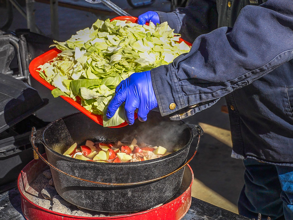 adding cabbage to ingredients in cast iron pot over hot coals