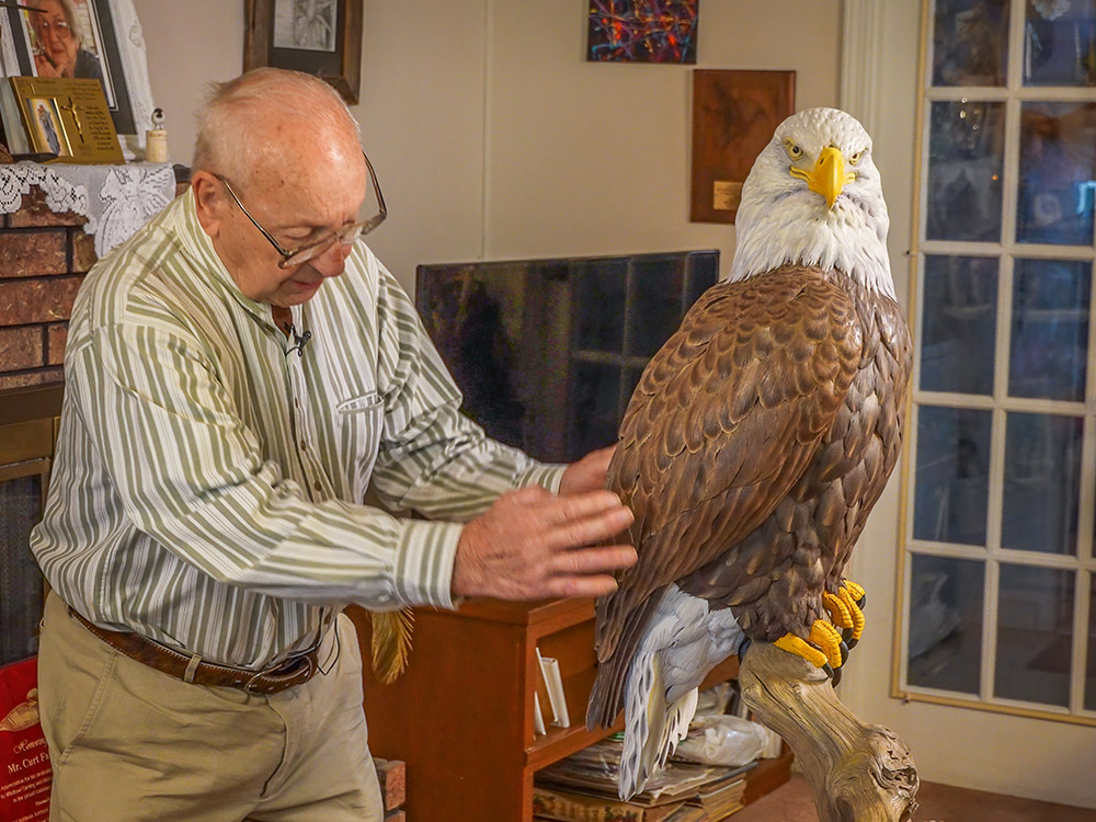 man dressed in long sleeved shirt with glasses and bald head places hands on wood bald eagle he carved