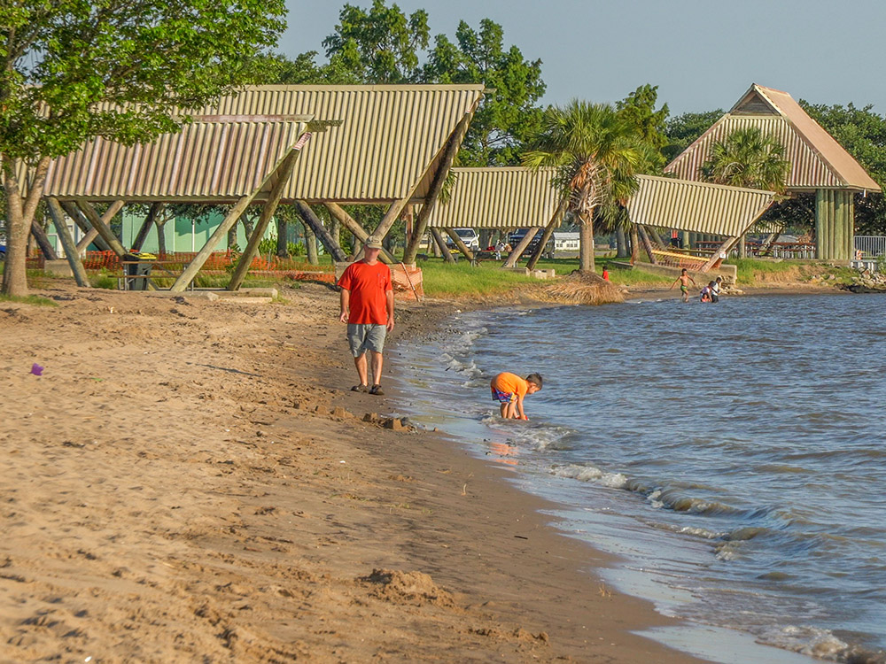 man in red shirt walks along beach while young child plays in water with covered pavilions trees in background