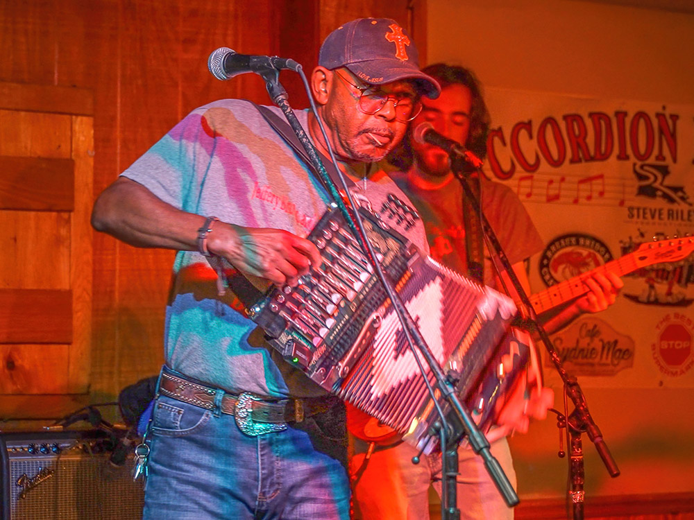 man wearing cap plays zydeco accordion on stage with guitar player in background
