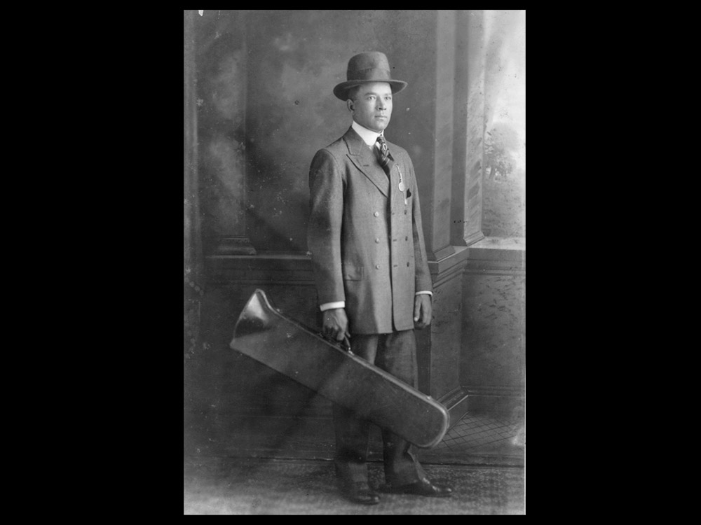 Kid Ory wearing suit and hat holding trombone case