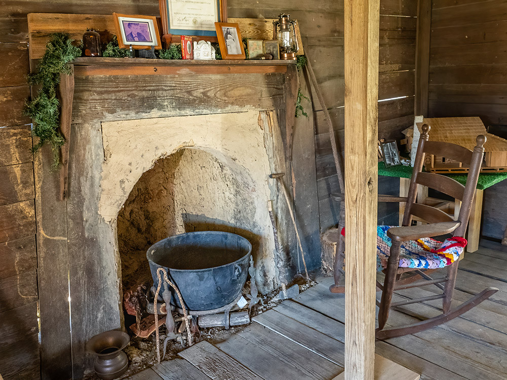 mud fireplace and black iron kettle and rocking chair inside log cabin