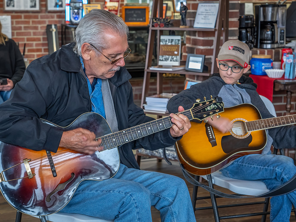 older man plays acoustic guitar along with younger boy on guitar wearing cap at bluegradd jam
