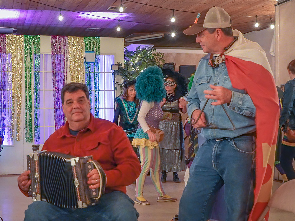 seated man in red shirt plays accordion and man standing in cap and blue jeans plays triangle at Cajun music jam in Thibodaux