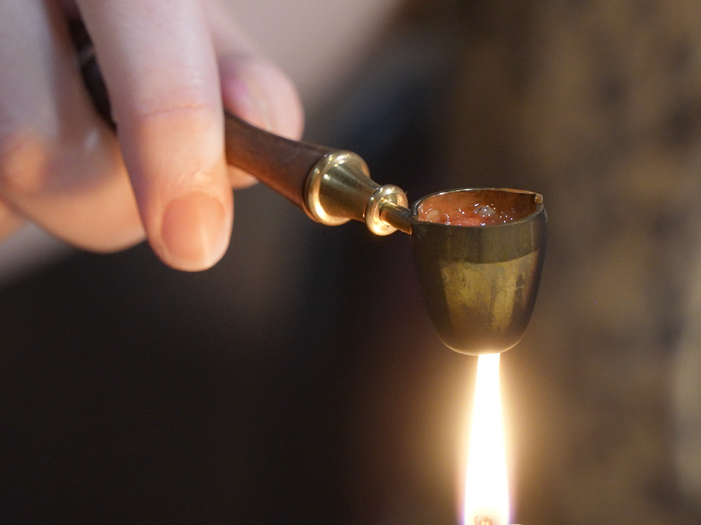 hand holding small brass cup over candle flame to melt wax