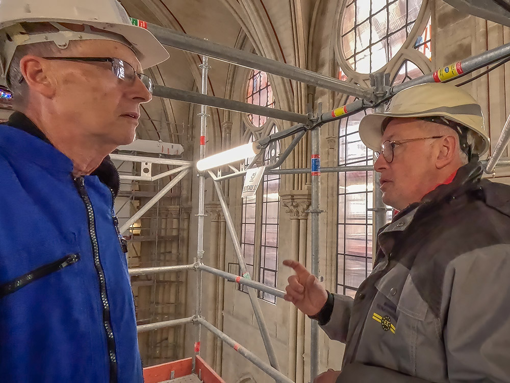 man in blue clothing and white hard hat with man in dark gray jacket and white hard hat in scaffolding in Notre Dame Cathedral