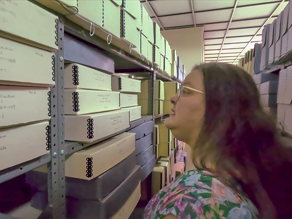 woman wearing glasses with brown hair and print dress looks over boxes on library shelves