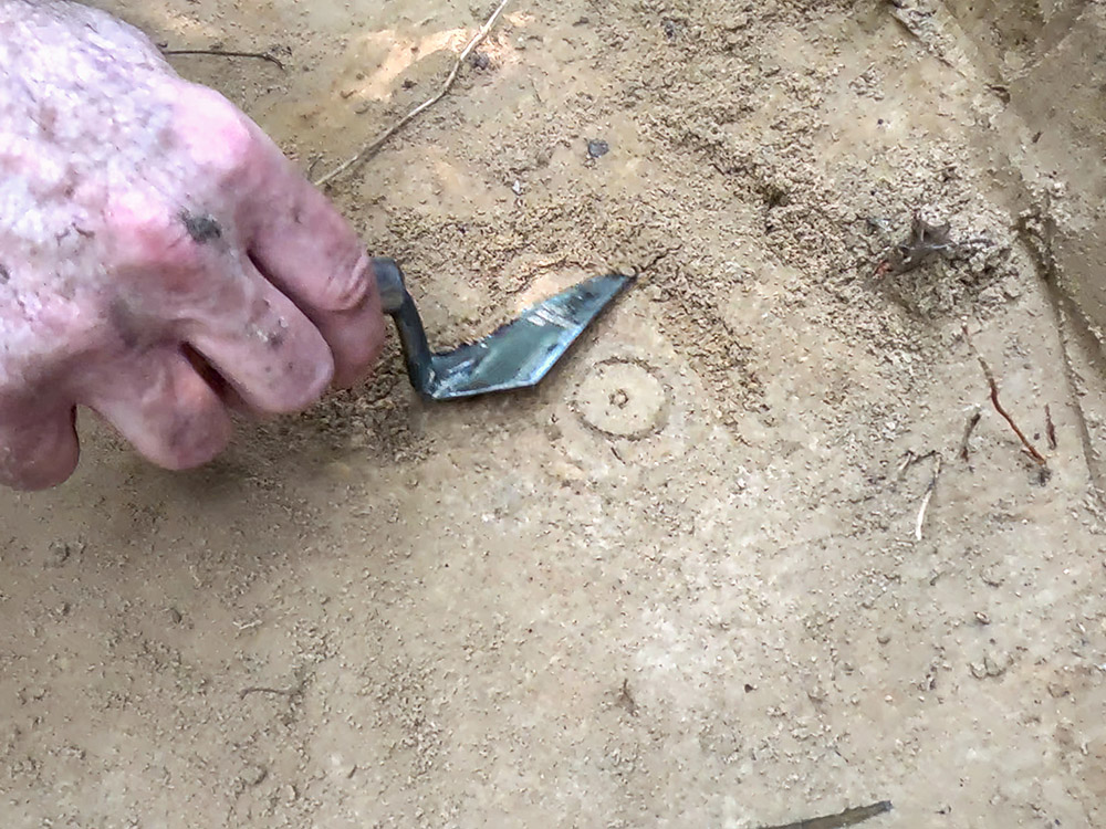hand holding small metal scraper removing dirt from small circular feature in the dirt