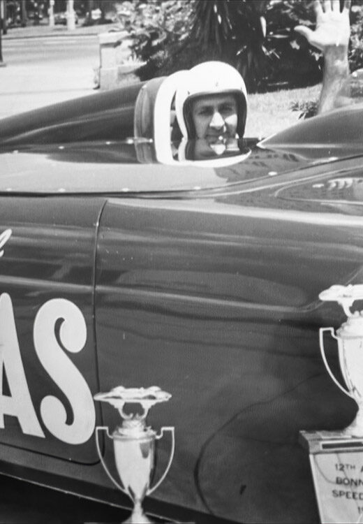 black and white photo of man in race car with trophy waving at camera