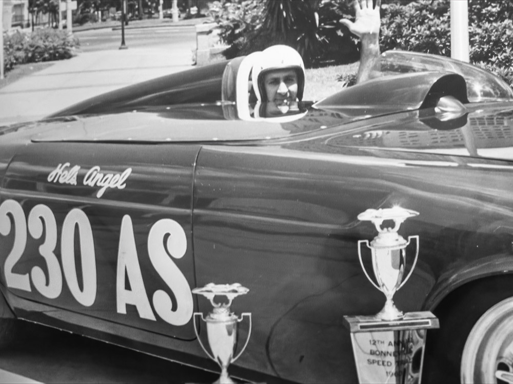 black and white photo of man in race car with trophy waving at camera