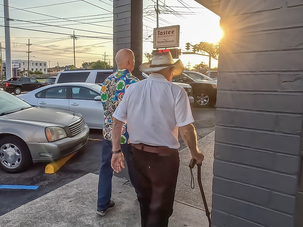man in white shirt with cane and straw hat and friend on sidewalk with morning sun