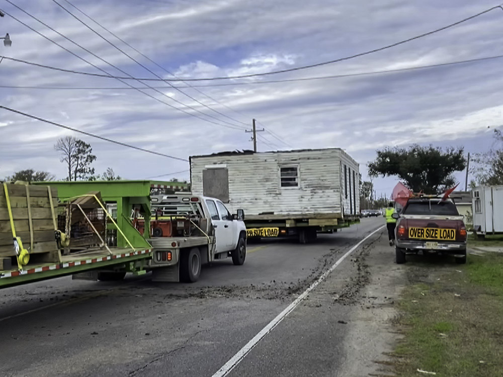 white wooden house being moved on trailer along highway with escort
