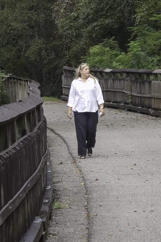 woman with blond hair, white shirt and black pants walks along footbridge in forest