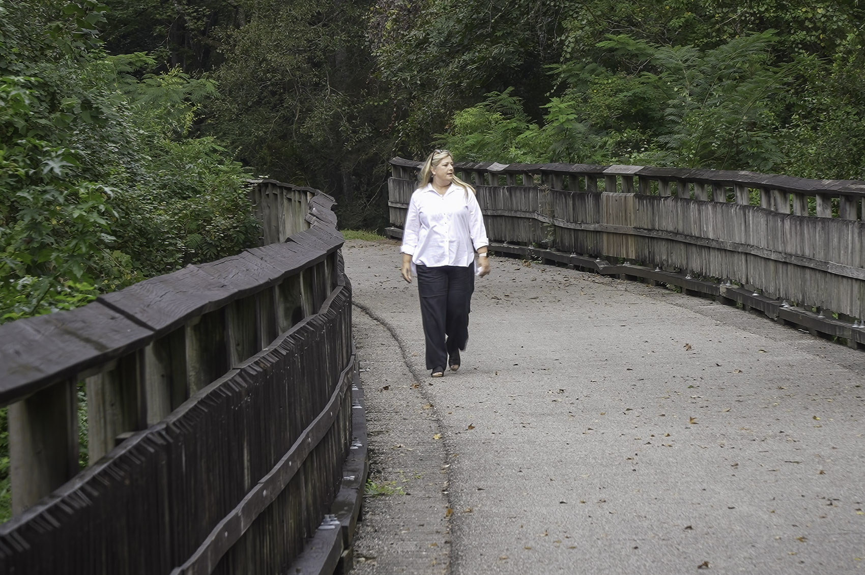 woman with blond hair, white shirt and black pants walks along footbridge in forest