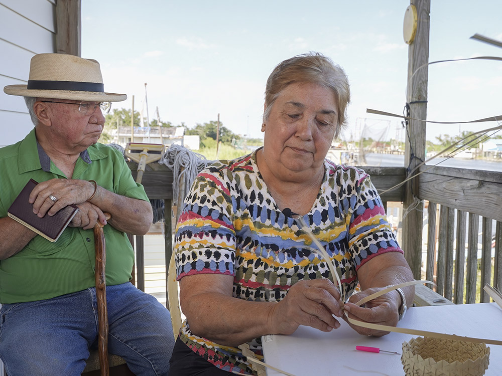 man with cane with white hat, green shirt and blue jeans sits next to woman making basket at table