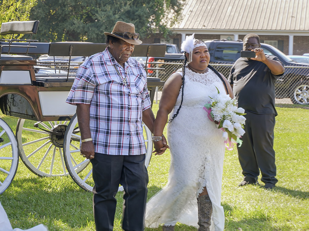  man in plaid shirt and hat escorts woman in white wedding dress
