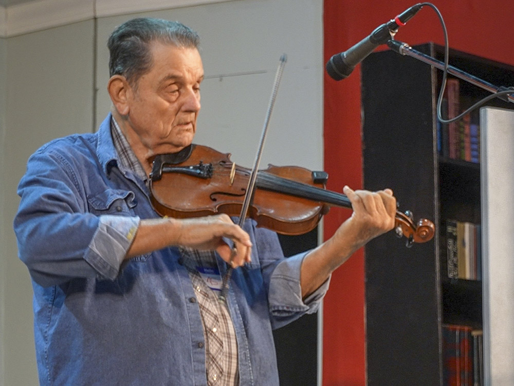 elderly fiddler in blue shirt on stage in front of microphone