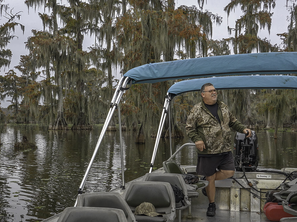  man in camouflage colored jacket guide tour boat through cypress trees with fall color.