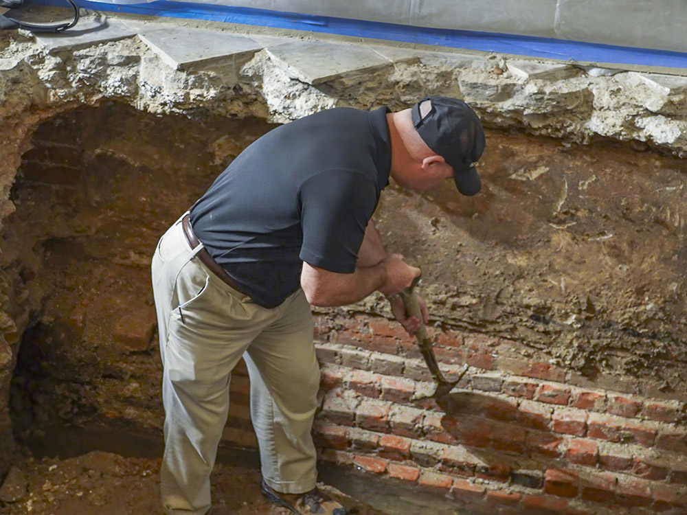 man wearing cap, black shirt, tan pants with shovel taps brick foundation
