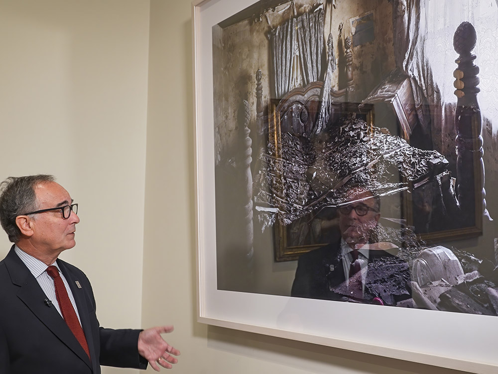 man in dark suit, red tie and glasses stands in front of large image of damaged home handing on wall