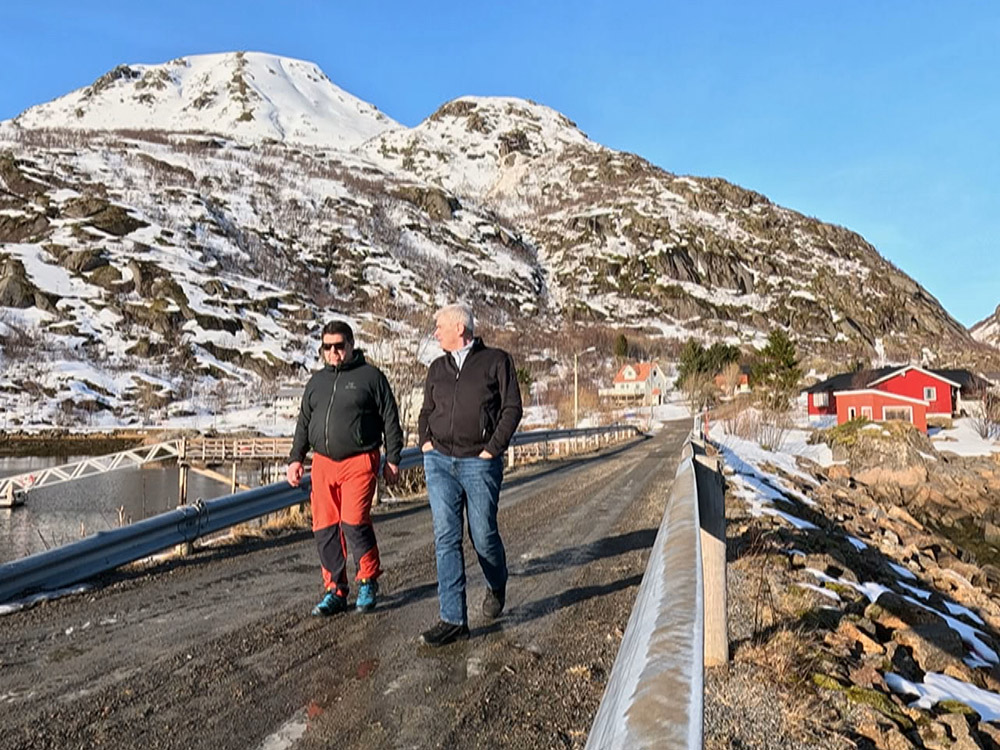 2 men walking along wet gravel road with snow covered mountainin background under blue sky