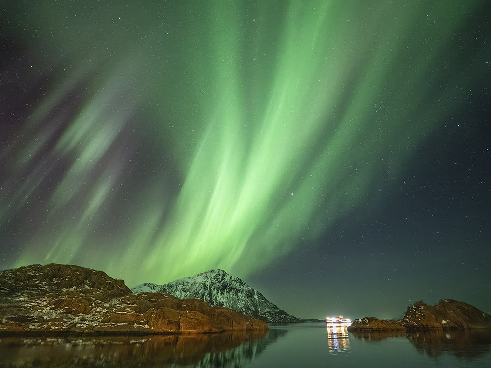 ferry with lights passing mountains underneath streaking northern lights
