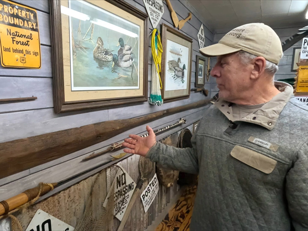 man with tan cap and gray jacket points to old wooden fishing oar hanging on wall