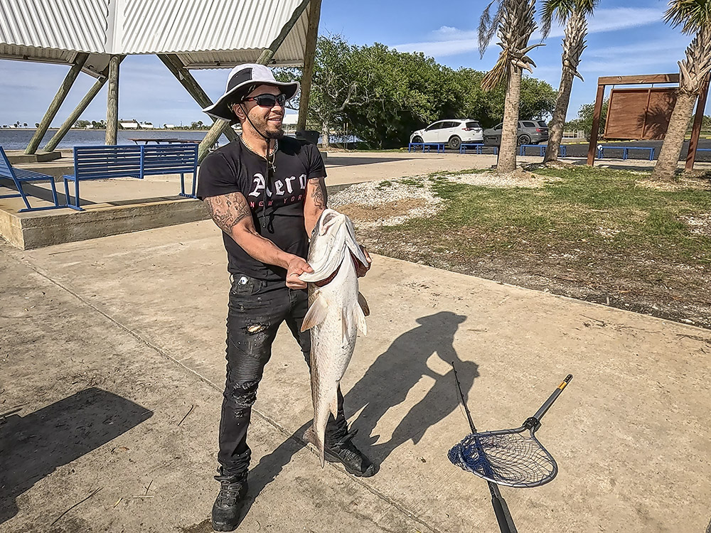 man in black shirt jeans and hat holding up large bull redfish