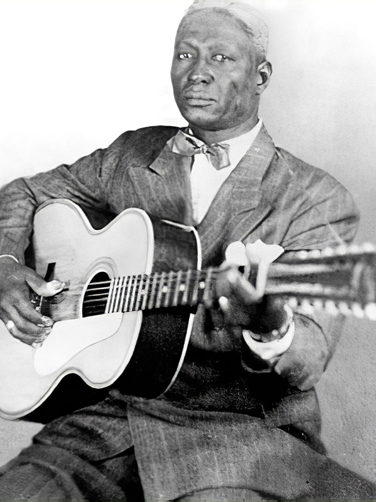 black and white photograph of man in suit playing 12 string guitar.