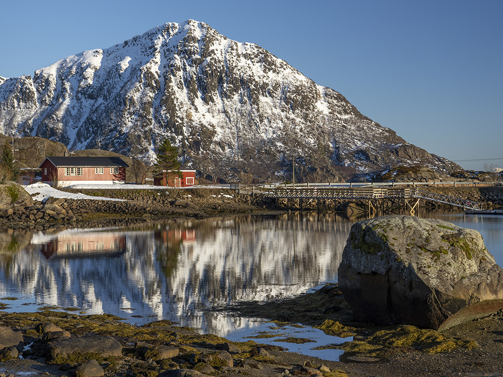 snow capped mountain reflected in calm pool of water with red painted houses and pier under blue sky in Norway