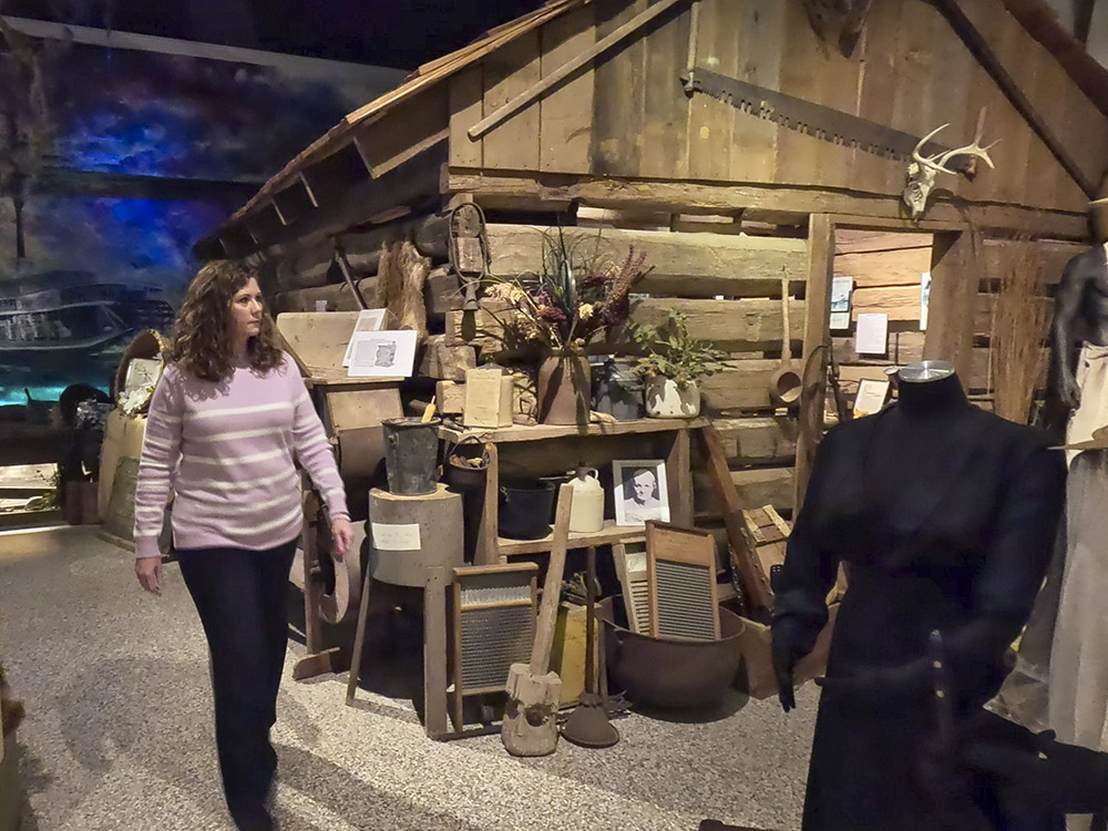woman with brown hair striped lavender sweater and dark pants walks past log cabin in museum