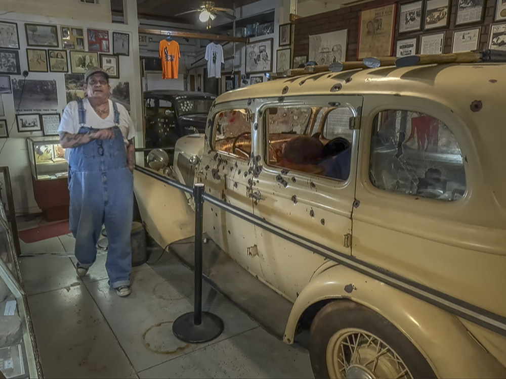 man in white shirt and blue coveralls walks past antique yellow car with bullet holes in museum