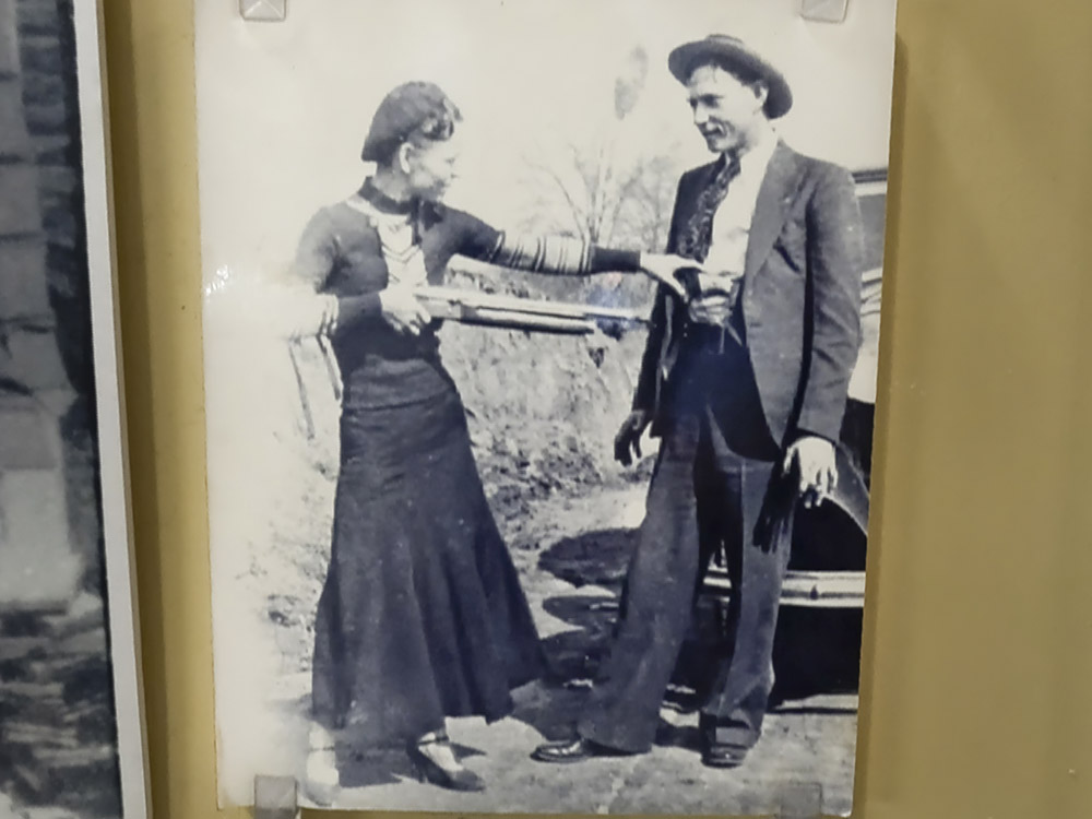photo of bonnie and clyde with woman holding shotgun pointed at man in front of car