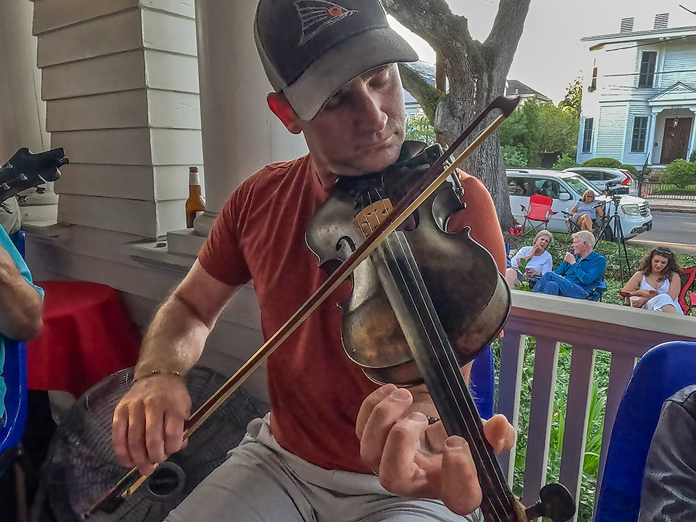 man wearing cap and orange shirt plays fiddle on porch