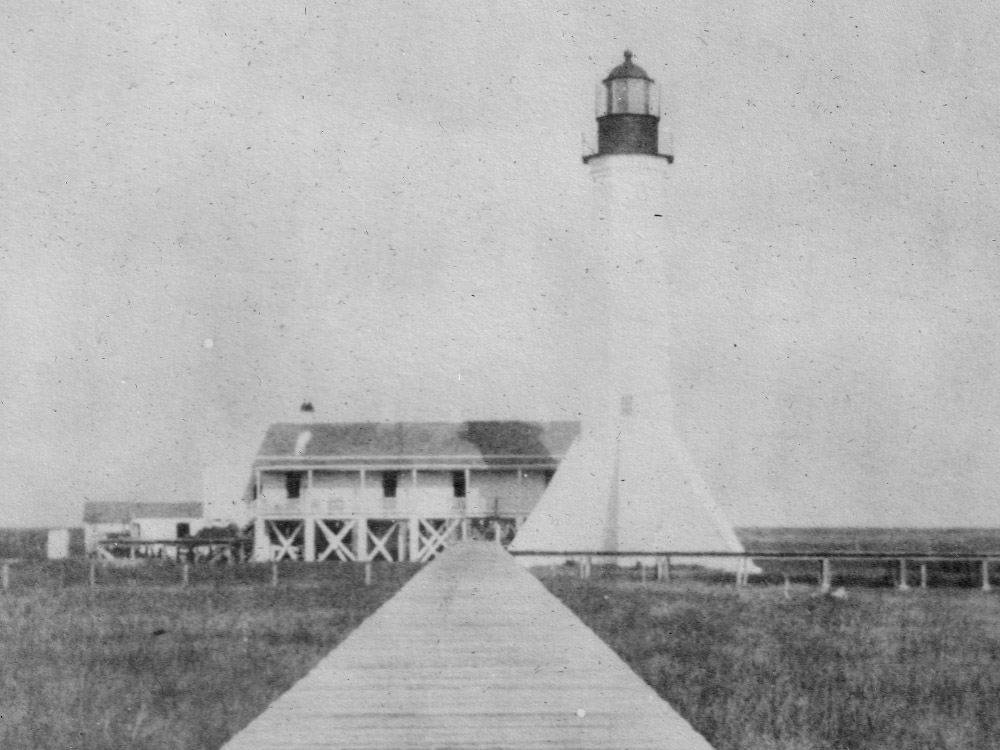 black and white picture of white brick lighthouse with black metal top and raised white wooden house