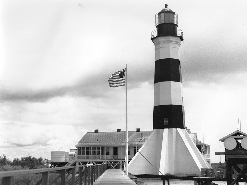 black and white image of brick lighthouse with black and white horizontal stripes, white building and u.s. flag