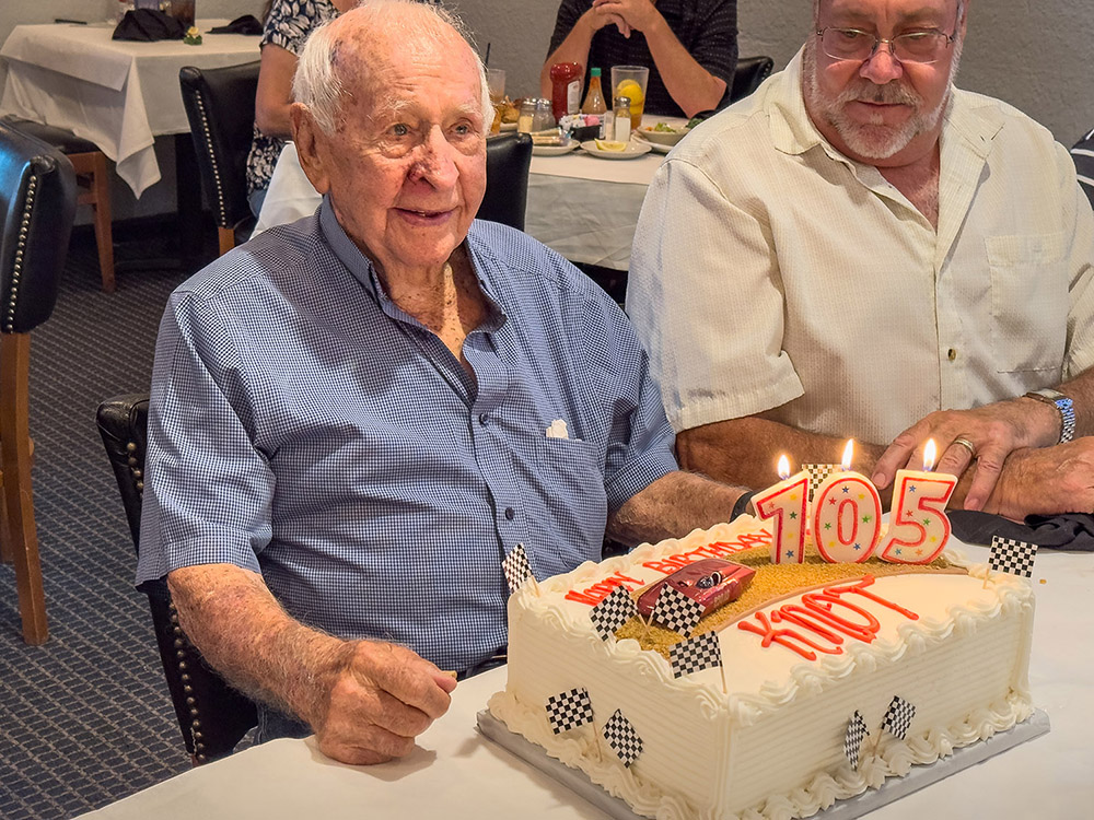 man with balding gray hair wearing blue shirt sits in front of birthday cake as he turns 105