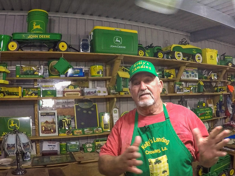 man wearing green hat, red shirt and green apron in front of toy tractors on shelves