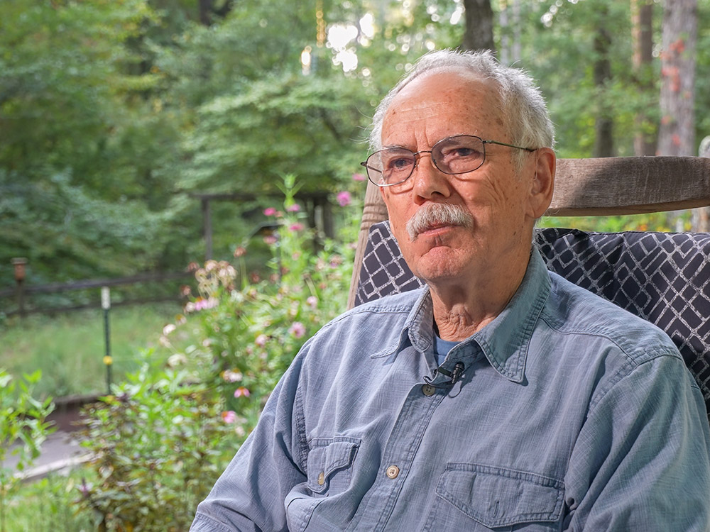 man with gray hair and moustache and blue shirt sitting in chair on porch