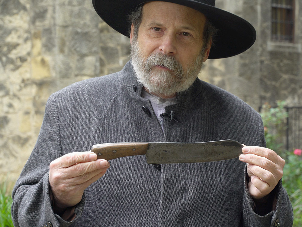 man in black hat and gray coat holds a Bowie knife