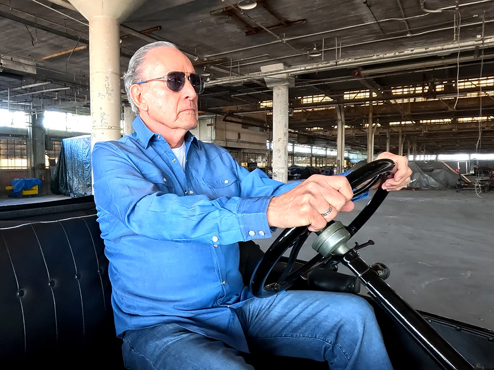 man wearing blue shirt and jeans and sunglasses sits in drivers seat of model T in Ford Assembly Plant