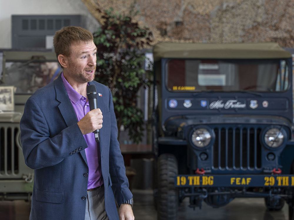 man with short hair and beard purple shirt and blue jacked holding microphone near military jeep