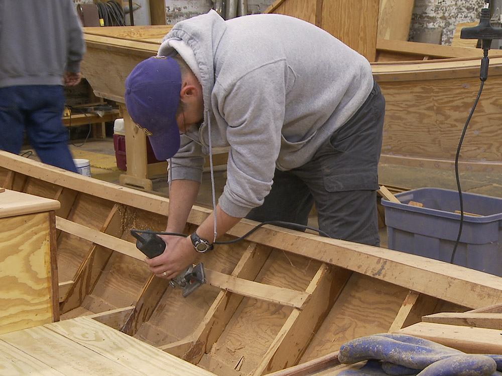 man wearing purple hat and gray sweatshirt sanding traditional Cajun boat