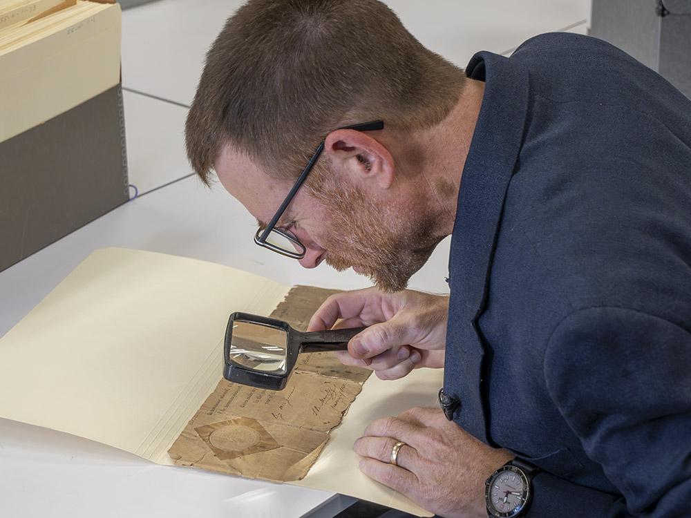 man with glasses, magnifying glass, wearing blue jacket looking at old document 