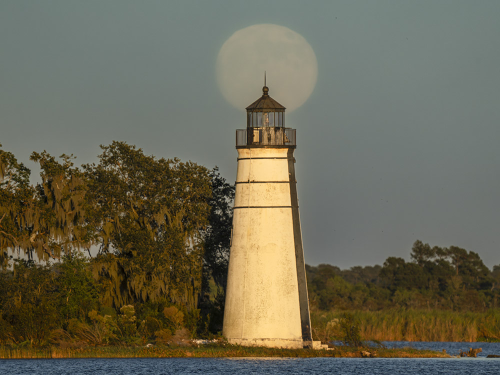 supermoon rises behind lighthouse in golden daylight of setting sun