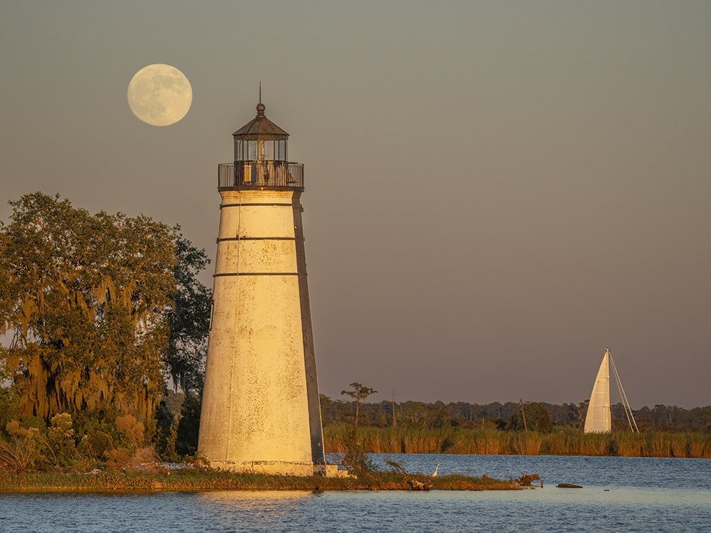 full moon near lighthouse on lake with sailboat in the background at sunset